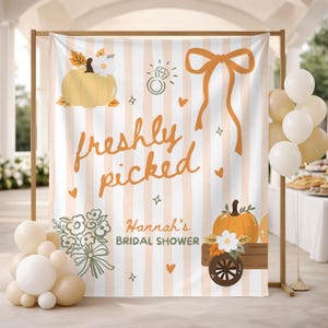 May include: A rectangular fabric backdrop featuring vertical light orange and white stripes. The text reads "freshly picked" in orange script and "Hannah's BRIDAL SHOWER" in dark green. Graphics include pumpkins, a ring, a bow, and a flower bouquet.