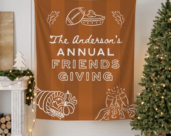 Op de afbeelding: Een bruine wandtapijt met witte illustraties en tekst. De tekst luidt "The Anderson's ANNUAL FRIENDS GIVING". Illustraties zijn onder meer een voetbal, taart, hoorn des overvloeds, bladeren en champagneglazen. Een kerstboom en een open haard staan op de achtergrond.