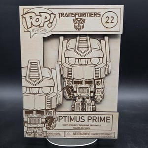 May include: A laser-cut wooden Optimus Prime figure in a display box. The box features the Pop! Customs and Transformers logos. The figure is intricately detailed. The text "OPTIMUS PRIME" is visible on the box.