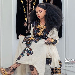 Puede incluir: Una mujer con un vestido blanco bordado con detalles negros y dorados. Lleva el pelo en dos trenzas y sostiene un trozo de tela en sus manos. El vestido tiene un diseño tradicional etíope.