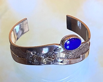 May include: A silver bracelet with a blue stone set in the center. The bracelet is decorated with a detailed design of a bird in flight.