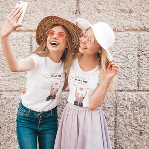 May include: Two friends wearing white t-shirts with the text "BRIDESMAIDS MAKE THE memories" and a graphic of two women holding hands. One friend is wearing a straw hat and sunglasses, the other is wearing a white sun hat and sunglasses. They are standing in front of a stone wall and taking a selfie.