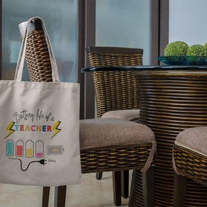 May include: A beige tote bag with the text "Battery life of a TEACHER" and a battery graphic. The graphic shows a series of batteries in various colors and states of charge, with a plug at the end. The bag is hanging in a room.