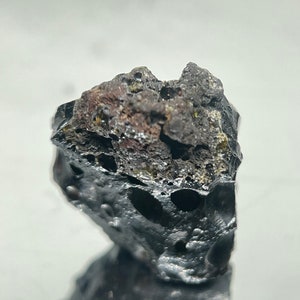May include: A dark gray, rough, and porous rock with a slightly metallic sheen. The rock has a unique, irregular shape and appears to be a meteorite.