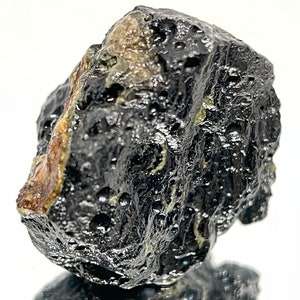 May include: A dark gray, rough-textured meteorite with a small patch of lighter gray and brown. The meteorite is about the size of a golf ball.