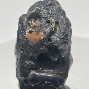 May include: A dark gray volcanic rock with a rough, textured surface. The rock has a large, irregular hole in the center, and smaller holes and indentations throughout. The rock has a reddish-brown color in the center of the hole.