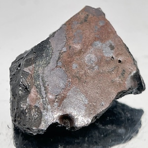 May include: A close-up of a dark gray, metallic rock with a rough, uneven surface. The rock has a mottled appearance with patches of silver and brown.