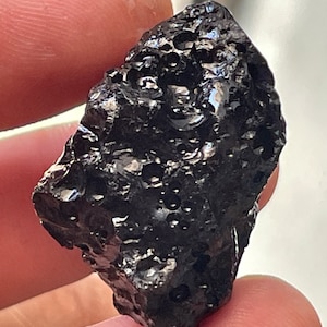 May include: A black, rough, and porous rock with a shiny, glassy surface. The rock has a unique, irregular shape and appears to be volcanic in origin.