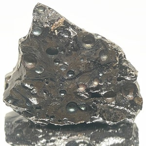 May include: A dark gray volcanic rock with a rough, textured surface. The rock has many small, round, black and iridescent bubbles embedded in it.