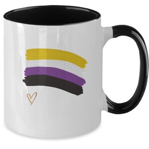 Puede incluir: Taza de cerámica blanca con interior y asa negros. La taza presenta un diseño de bandera del orgullo no binario en pinceladas amarillas, blancas, moradas y negras, con un pequeño corazón dorado. Una taza de café sencilla y elegante.