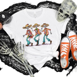 May include: White t-shirt with a graphic of three skeletons wearing sombreros and playing music. The skeletons are wearing colorful clothing and have a festive look.
