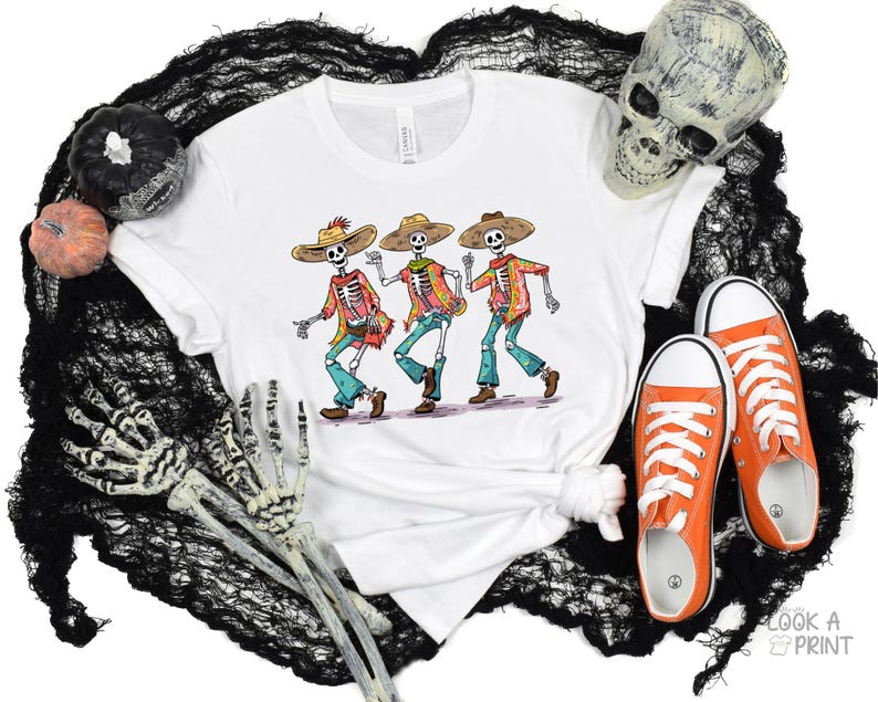 May include: White t-shirt with a graphic of three skeletons wearing sombreros and playing music. The skeletons are wearing colorful clothing and have a festive look.