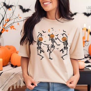 May include: A beige t-shirt with a graphic of three skeletons dancing with pumpkins for heads. The skeletons are wearing a black outline and are dancing in a circle. The background is a light beige color with a few bats and stars.