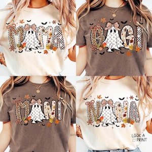 Puede incluir: Cuatro camisetas con diseños temáticos de otoño. Cada camiseta presenta una palabra diferente: "MAMA", "MAGI", "MANI" y "NANA". Los diseños incluyen un fantasma, hojas de otoño y detalles de estampado de leopardo. Las camisetas son de varios colores neutros.
