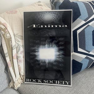 May include: A framed black and white poster with the word "Enigma" in white lettering at the top. The poster has a bright white square in the center and the words "ROCK SOCIETY" at the bottom.
