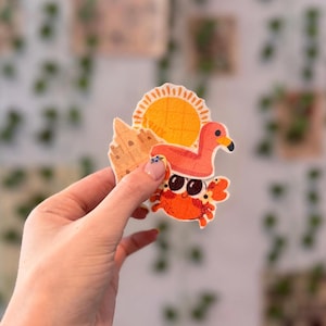 May include: A collection of colorful, beach-themed stickers. The stickers feature a crab wearing sunglasses, a flamingo, a sun, and a sandcastle. The stickers are held in a hand against a blurred background.