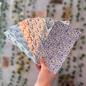 May include: A hand holds a fanned stack of patterned paper. The papers feature nautical designs in shades of blue, orange, and white. Designs include waves, coral, and nautical symbols. The paper has a textured appearance.