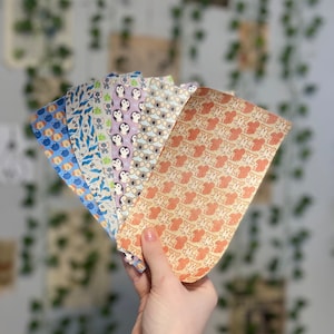 May include: A set of six colorful patterned paper sheets featuring various animal designs, including penguins, koalas, and dogs. The sheets are held in a hand.