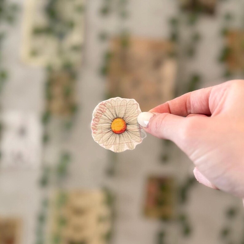May include: A white daisy flower sticker with a yellow center. The sticker is printed on a textured paper and has a slightly irregular shape.