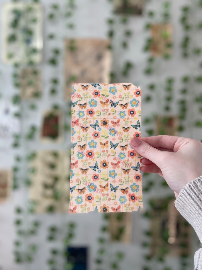 Pu&ograve; includere: Un foglio di carta rosa con un motivo ripetitivo di farfalle e fiori. Le farfalle sono blu, arancioni e gialle. I fiori sono rosa, blu e gialli.