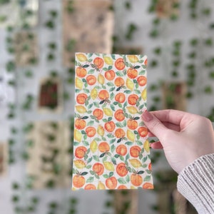 Pu&ograve; includere: Un foglio di tessuto bianco con un motivo ripetuto di pesche arancioni e limoni gialli con foglie verdi. Il tessuto ha un motivo a griglia.