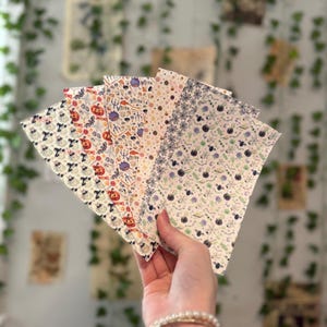 May include: A set of five rectangular sheets of paper arranged in a fan shape, each featuring a distinct Halloween-themed pattern including pumpkins, ghosts, witches, black cats, spider webs, cauldrons, and various small seasonal icons.