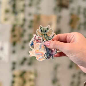 May include: A hand holds a collection of whimsical stickers. The stickers feature illustrations of a green dragon, a small village, a unicorn, and a vintage bicycle. The stickers have a watercolor style with soft pastel colors.