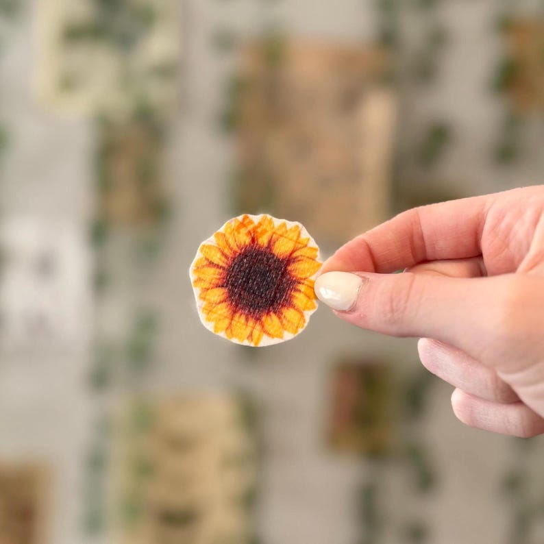 May include: A yellow and brown sunflower sticker with a white background.