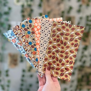 May include: A hand holds a fan of fabric pieces, each displaying a different sports-themed pattern. Designs include soccer balls, basketballs, footballs, and volleyballs in various colors. The fabrics have a light background.