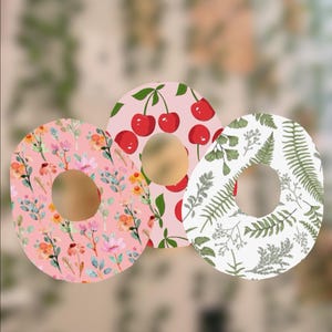 May include: Three decorative donut-shaped designs. One features pink flowers and green leaves on a pink background. Another has red cherries on a pink background. The third has green ferns on a white background.