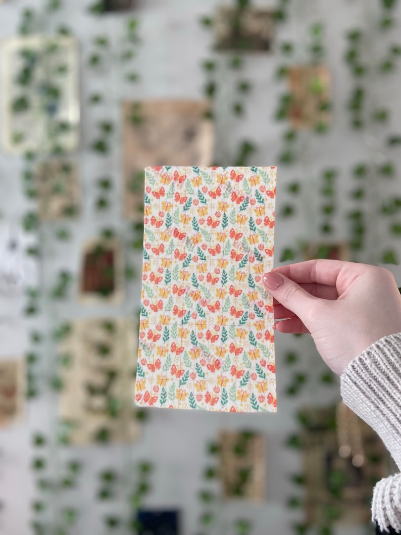 Pu&ograve; includere: Un tessuto bianco con un motivo ripetitivo di farfalle colorate e foglie verdi. Il tessuto &egrave; di forma quadrata ed &egrave; tenuto da una mano.