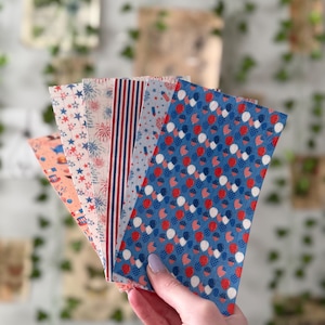 May include: A hand holds a fanned assortment of patterned paper sheets. The sheets feature patriotic designs with red, white, and blue colors, including stars, stripes, fireworks, and polka dots. The paper is likely for crafting or scrapbooking.