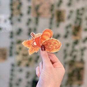 May include: A hand holds a collection of fall-themed stickers. The stickers include a pumpkin, a pie, a cup of coffee, and a fall leaf. The stickers are in shades of orange, brown, and red. The background is a wall with green vines.