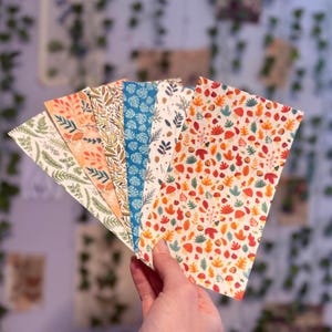 May include: A hand holds a fan of patterned paper sheets. The sheets display various botanical designs in colors like orange, green, and blue. The patterns include leaves, flowers, and other natural elements.