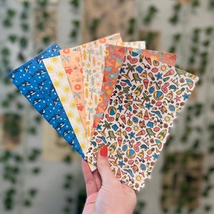 May include: A hand holds a fan of patterned papers with a scalloped edge. The papers feature summer-themed designs, including orcas, suns, surfboards, and beach elements in various colors. The background is blurred with greenery.