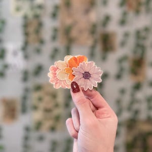 May include: A set of three paper flower stickers in shades of pink, orange, and white. The flowers are layered on top of each other and have a delicate, vintage look.