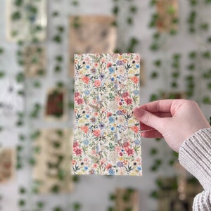 Pu&ograve; includere: Un foglio di tessuto con motivo floreale su sfondo bianco e fiori rosa, blu e gialli. Il foglio &egrave; tenuto da una mano.