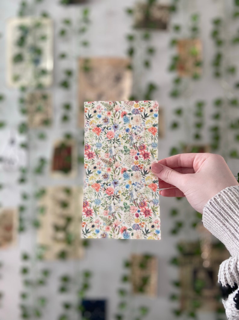 Pu&ograve; includere: Un foglio di tessuto con motivo floreale su sfondo bianco e fiori rosa, blu e gialli. Il foglio &egrave; tenuto da una mano.