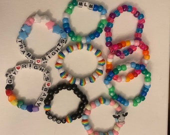 May include: A collection of colorful beaded bracelets with various words and symbols, including "LGBTQ+ Rights", "Gay", "Baby", "Crime", "MLM", and a cow charm.