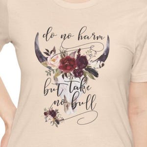 以下が含まれることがあります： ベージュのTシャツ。黒い筆記体で「do no harm but take no bull」の文字と、花のブルスカルのデザイン。フローラルアレンジメントには、バーガンディとピンクの花が含まれています。