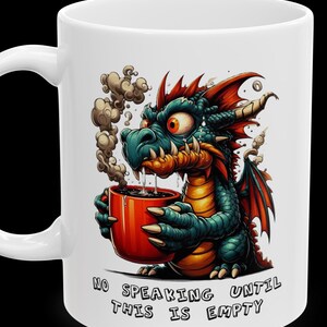 Op de afbeelding: Een witte keramische mok met een cartoon-draak die een rode mok koffie vasthoudt. De draak heeft groene schubben, rode vleugels en een chagrijnige uitdrukking. De tekst op de mok luidt "Niet praten tot deze leeg is."