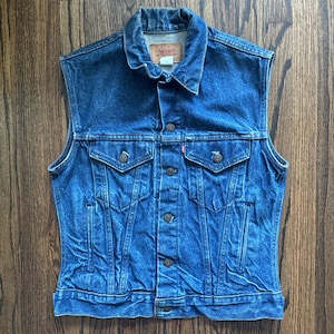 Può includere: Un giubbotto di jeans blu con chiusura a bottoni, due tasche sul petto e due tasche anteriori. Il giubbotto ha un'etichetta Levi's sulla tasca.