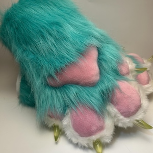 May include: A furry, teal-colored paw with pink pads and white fur accents. The paw has three translucent yellow claws. The item is likely a costume piece or part of a larger furry suit.