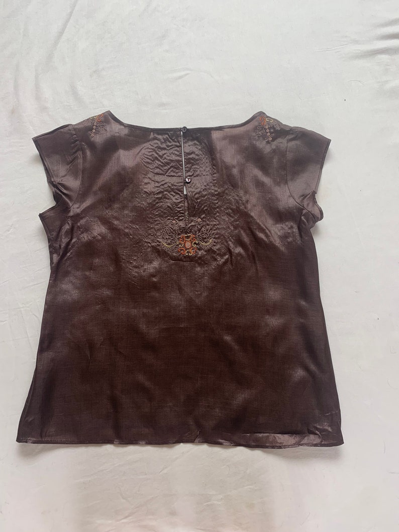 Puede incluir: Una blusa de seda marr&oacute;n con mangas cortas y detalles bordados. La blusa tiene un escote redondo y un cierre de bot&oacute;n en la espalda.