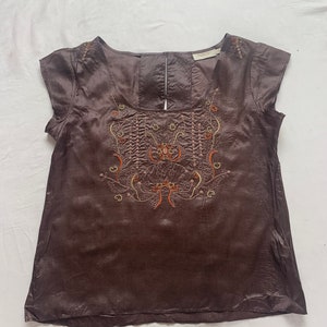 Puede incluir: Una blusa de seda marr&oacute;n con mangas cortas y un dise&ntilde;o floral bordado en la parte delantera. El bordado est&aacute; en tonos de naranja, amarillo y verde.