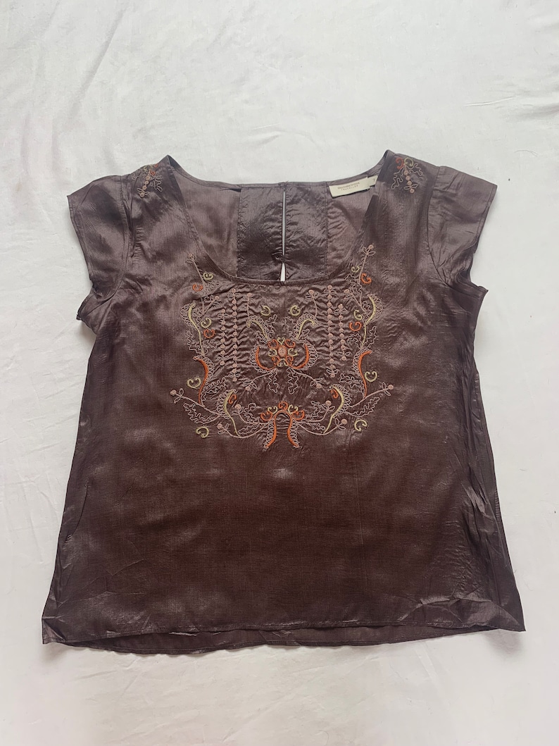 Puede incluir: Una blusa de seda marr&oacute;n con mangas cortas y un dise&ntilde;o floral bordado en la parte delantera. El bordado est&aacute; en tonos de naranja, amarillo y verde.