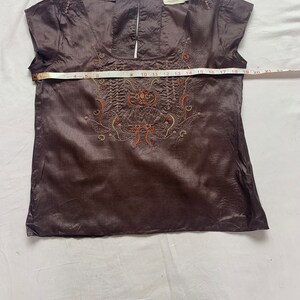 Puede incluir: Una blusa de seda marr&oacute;n con mangas cortas y un dise&ntilde;o floral bordado en la parte delantera. La blusa tiene un escote redondo y una abertura de ojo de cerradura en la espalda.