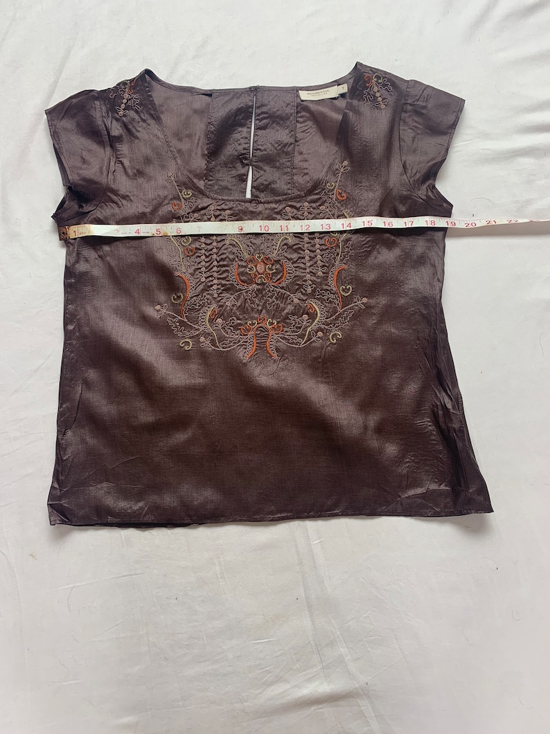 Puede incluir: Una blusa de seda marr&oacute;n con mangas cortas y un dise&ntilde;o floral bordado en la parte delantera. La blusa tiene un escote redondo y una abertura de ojo de cerradura en la espalda.