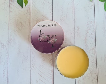 May include: A purple and white tin with the words "BEARD BALM" and a floral design. The tin is open and shows a yellow beard balm inside.