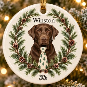 May include: A round white ceramic ornament featuring a chocolate Labrador retriever wearing a cream-colored tie with a green pine tree pattern. The dog is framed by a wreath of pine branches and pinecones, with the text Winston and 2026.